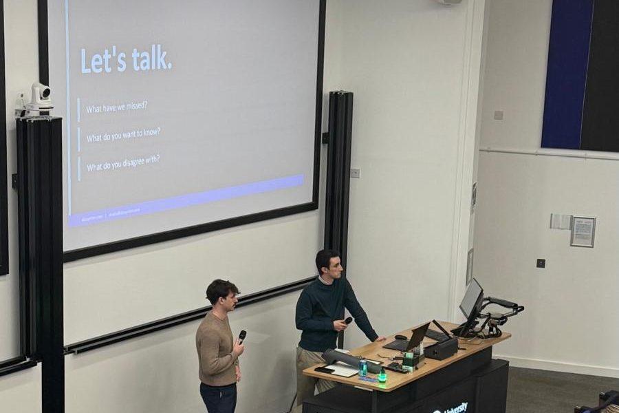 Charlie standing beside a lectern in a university lecture theatre holding a microphone, with a slide reading Let's talk on the screen behind him.