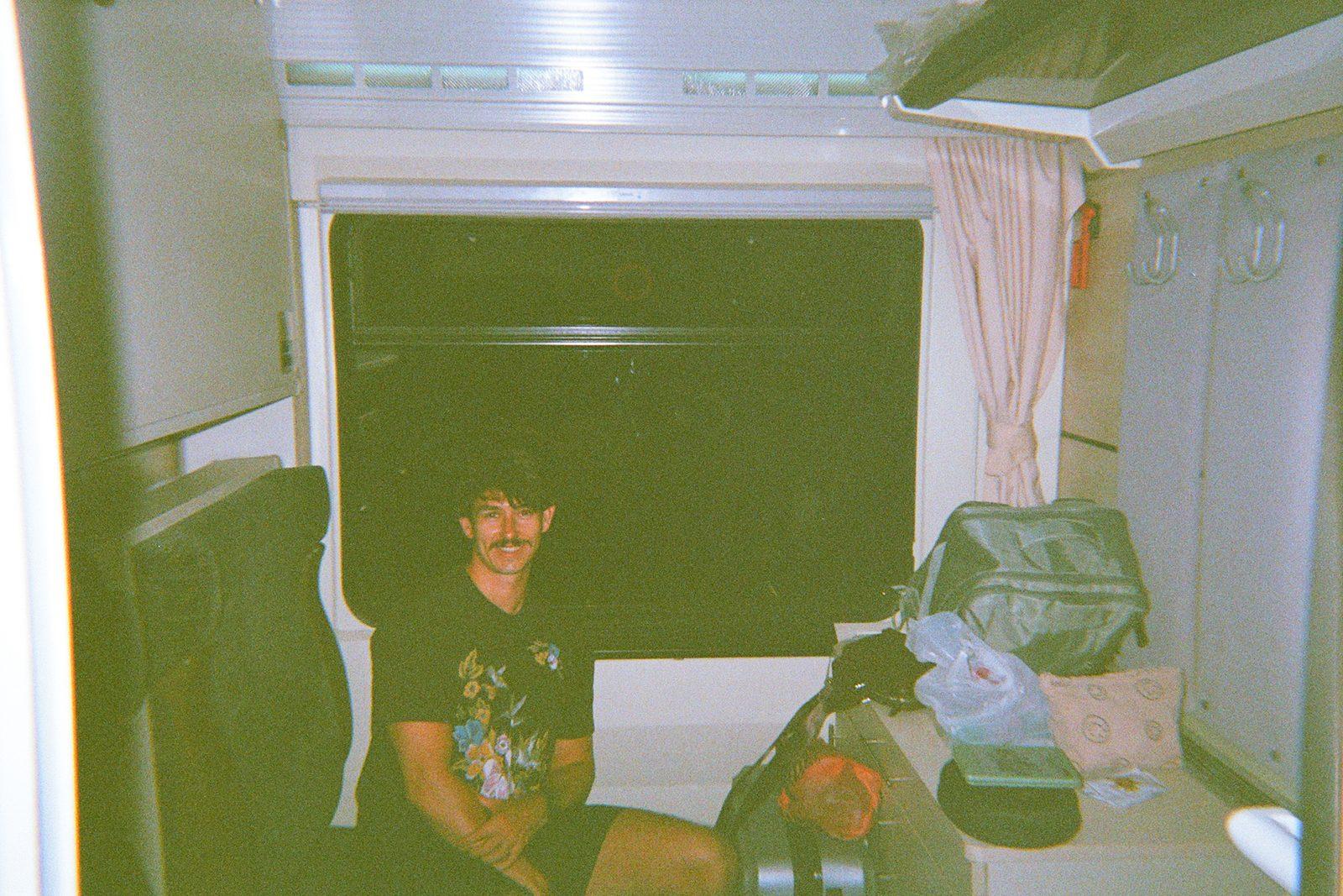 A green-tinted film photograph of Charlie in a floral black t-shirt sitting on a bunk in a sleeper train cabin, luggage stacked beside him.